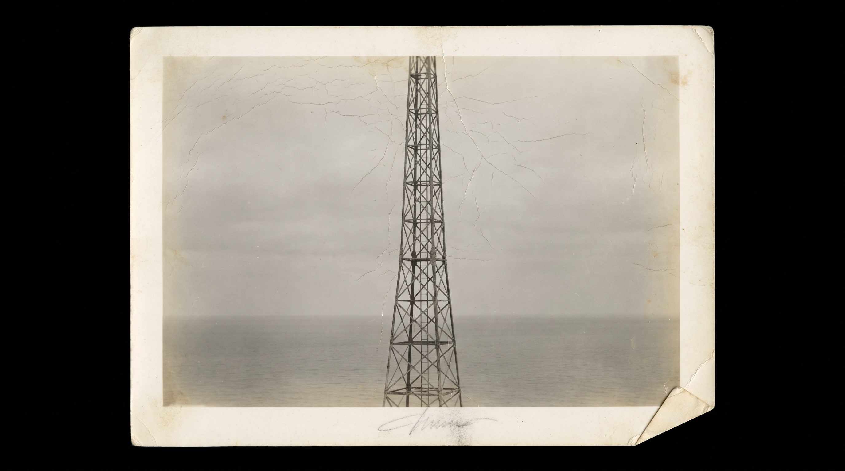 A black-and-white photograph of a coastal wireless mast against a flat grey sea, no figures present.